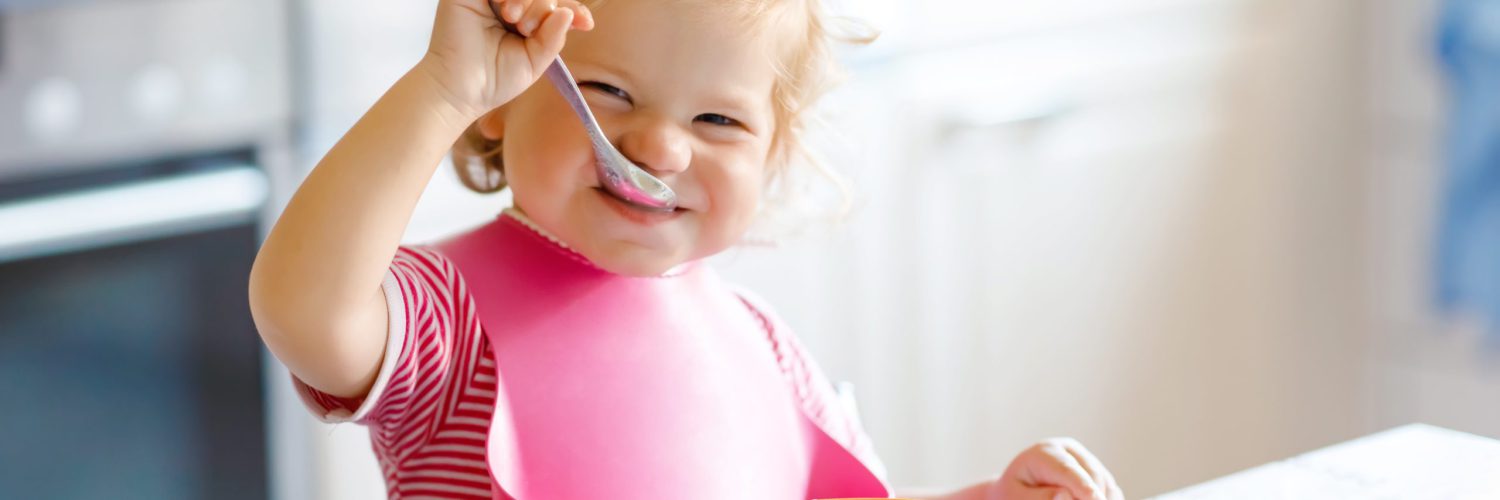 Adorable baby girl eating from spoon vegetable noodle soup. food, child, feeding and development concept. Cute toddler child, daughter with spoon sitting in highchair and learning to eat by itself.